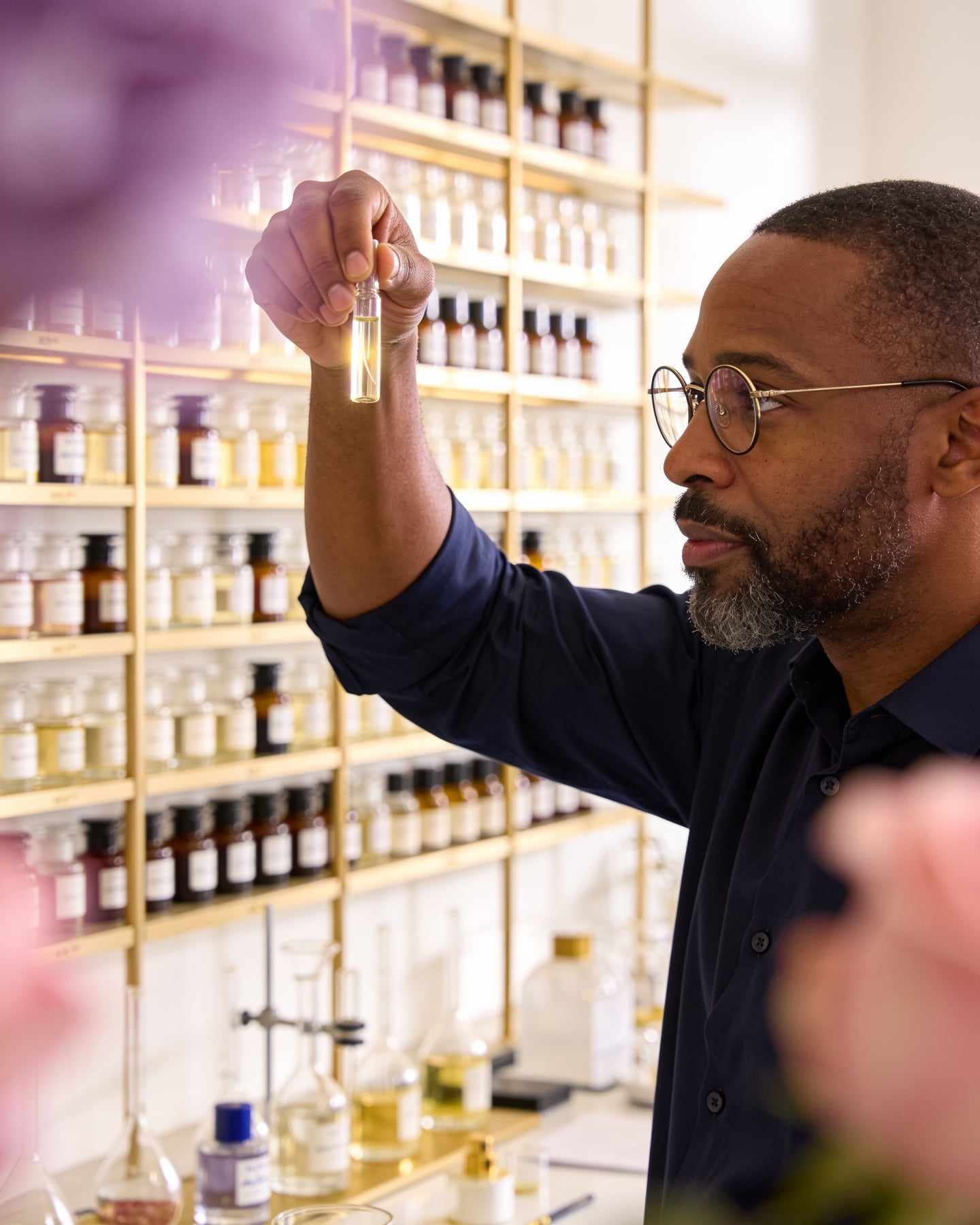 Portrait of a perfumer in his forties with a short beard and round glasses, holding a small glass vial up to the light.