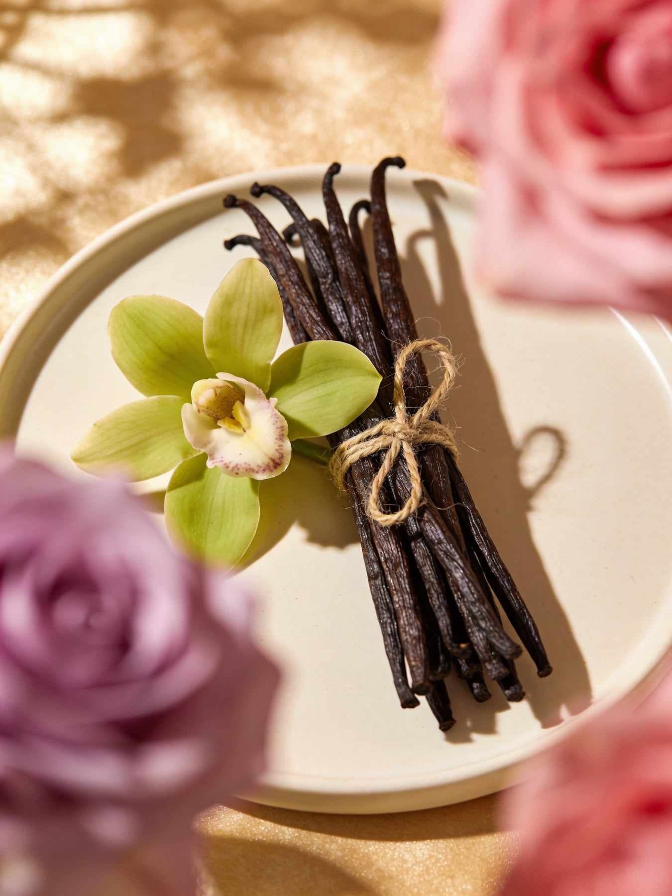 Cured dark vanilla pods tied with twine and a green vanilla orchid flower on a cream plate.